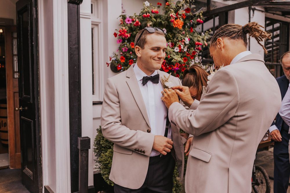 Wedding party members in beige suits assist with boutonniere pinning near red floral decorations.