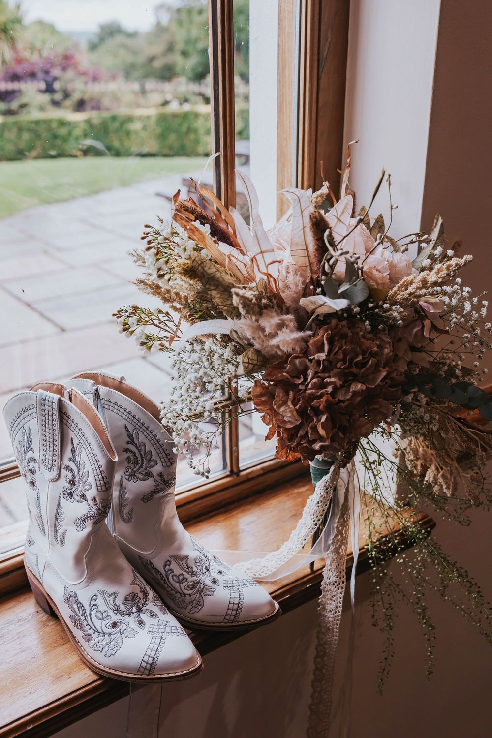 White cowboy boots and dried floral bouquet arranged on a rustic windowsill with garden view in soft natural light.