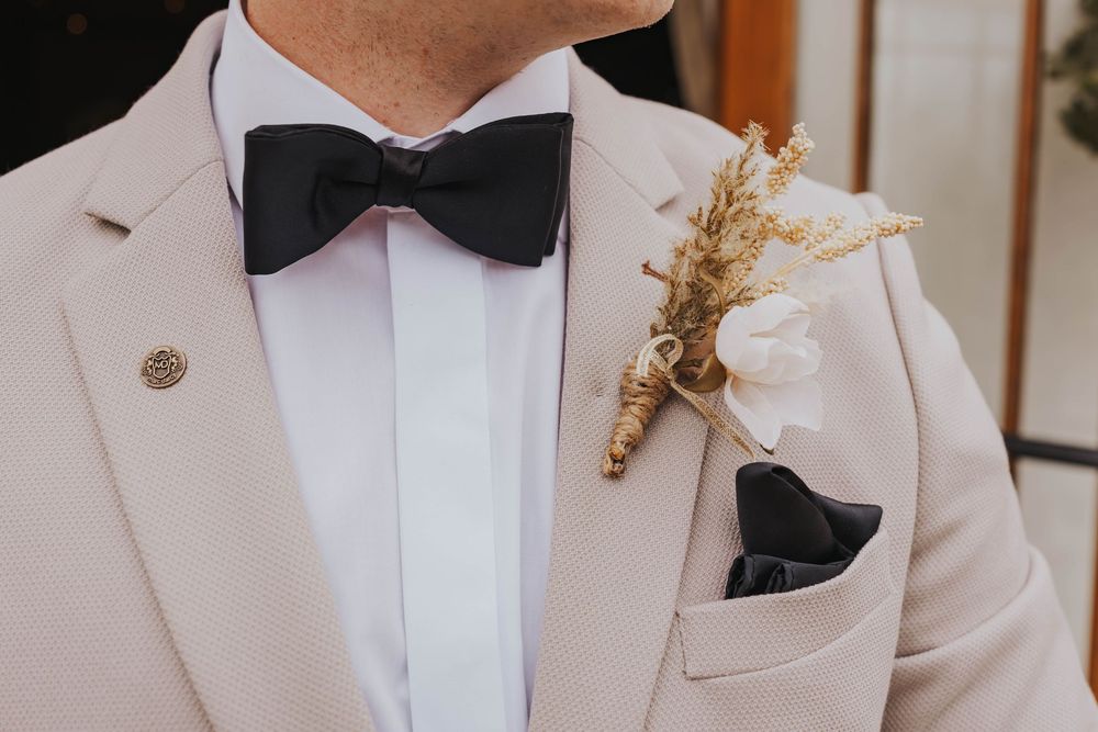 Elegant beige suit jacket with black bow tie and dried flower boutonniere detail.