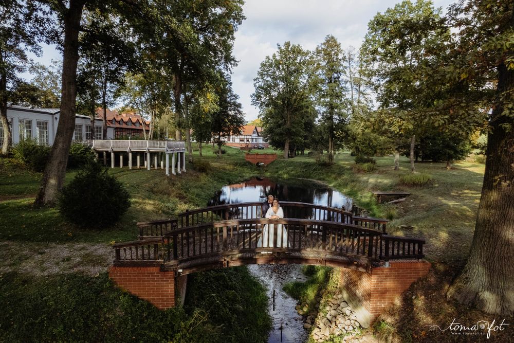 A wooden bridge spans over a reflective pond in a serene garden setting with trees and a white building in the background.
