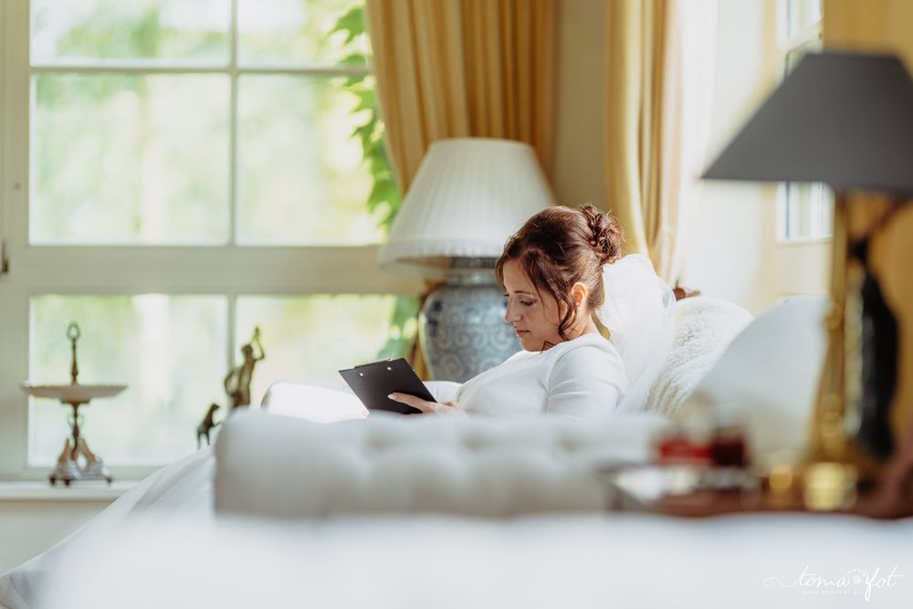 Someone relaxes while reading a tablet device in a bright, cozy bedroom with natural lighting and white bedding.