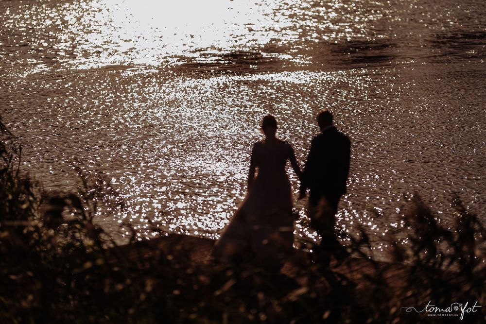 Two silhouetted figures stand along a glittering waterfront at sunset, creating a romantic atmospheric scene.