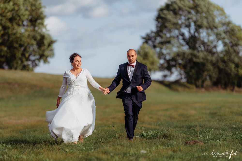 Joyful newlyweds run hand in hand across a green hillside under cloudy skies.