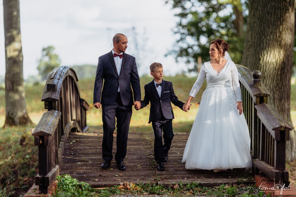 A newly married couple walks hand in hand with a young boy on a rustic wooden bridge surrounded by trees.