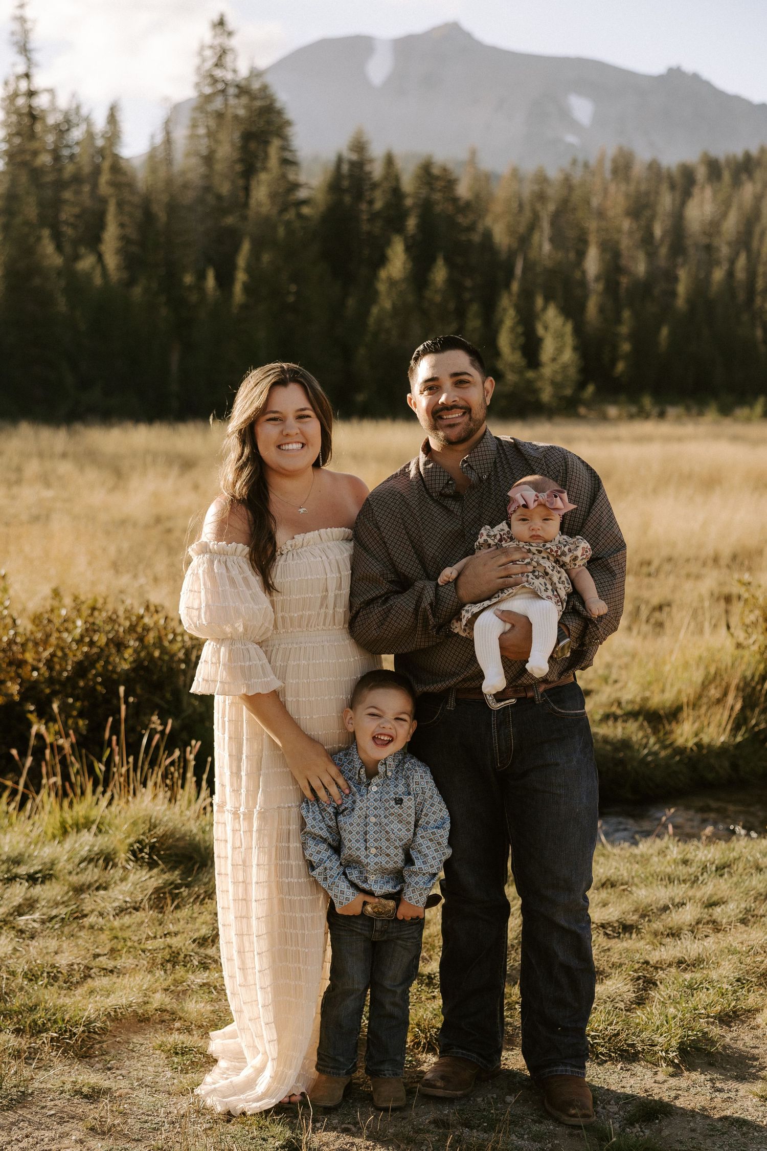 A family poses together in a scenic mountain meadow at sunset with evergreen trees in the background.
