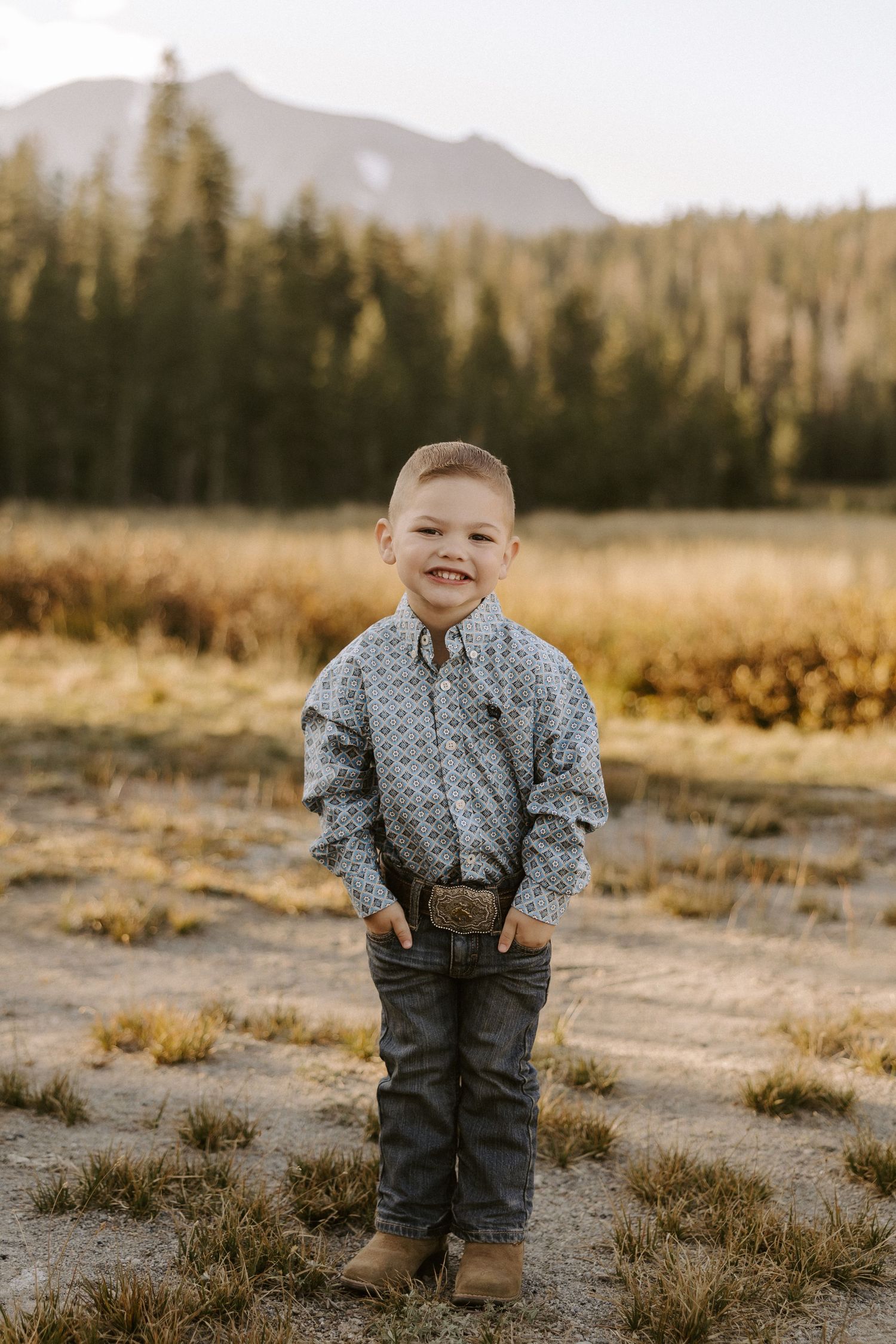 A young child stands outdoors in a button-up shirt and jeans against a scenic mountain and forest landscape.