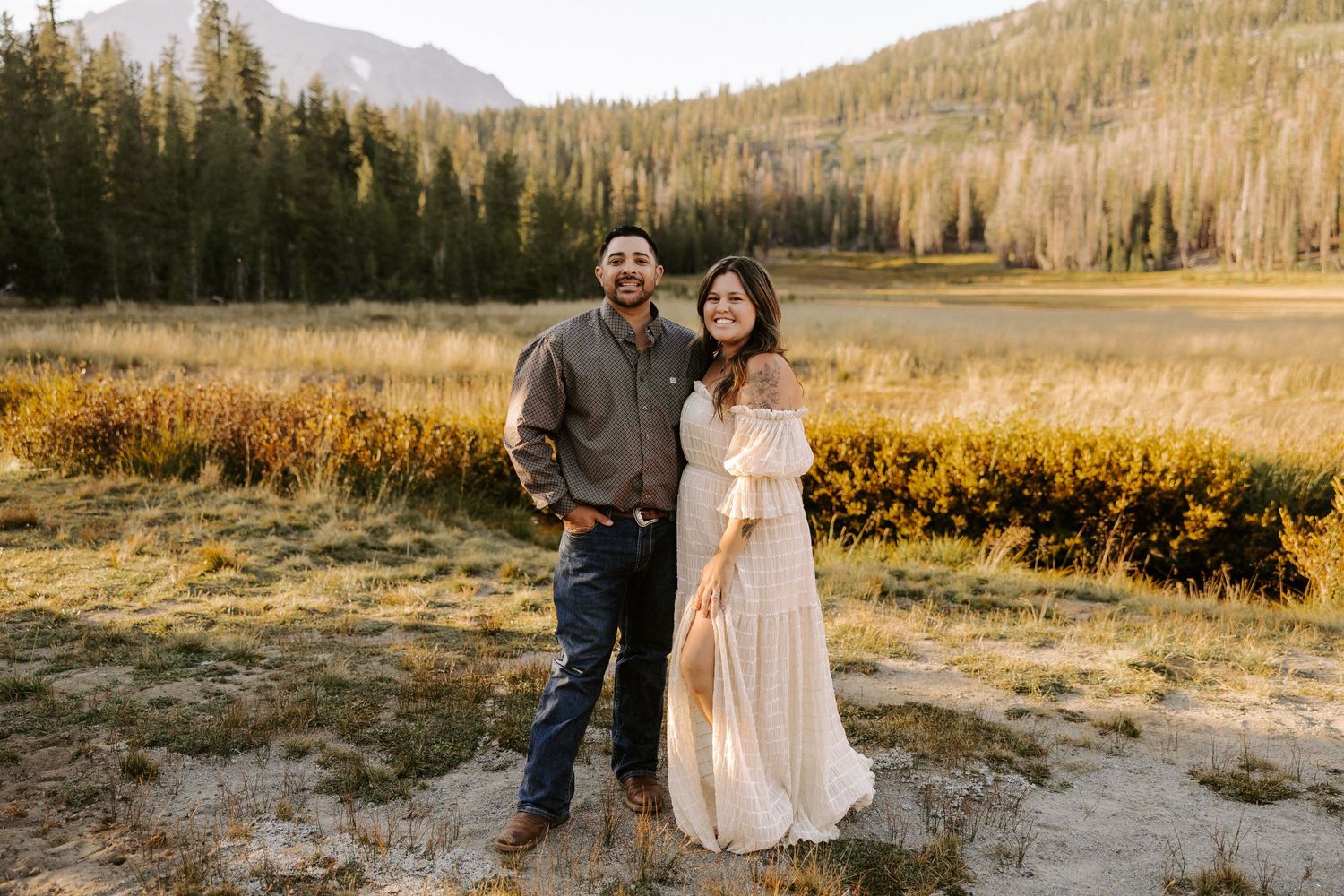 A couple stands together in a scenic mountain meadow during golden hour with evergreen forest and peaks in the background.