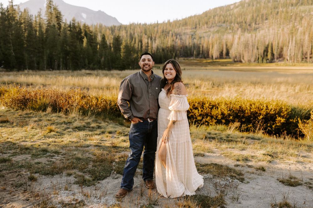 A couple stands together in a scenic mountain meadow during golden hour with evergreen forest and peaks in the background.