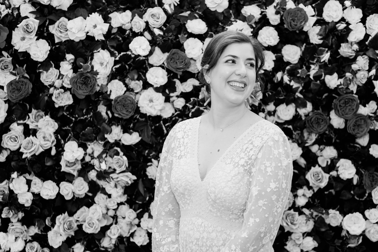 Black and white photo of a bride in a lace V-neck wedding dress smiling in front of a floral wall backdrop.