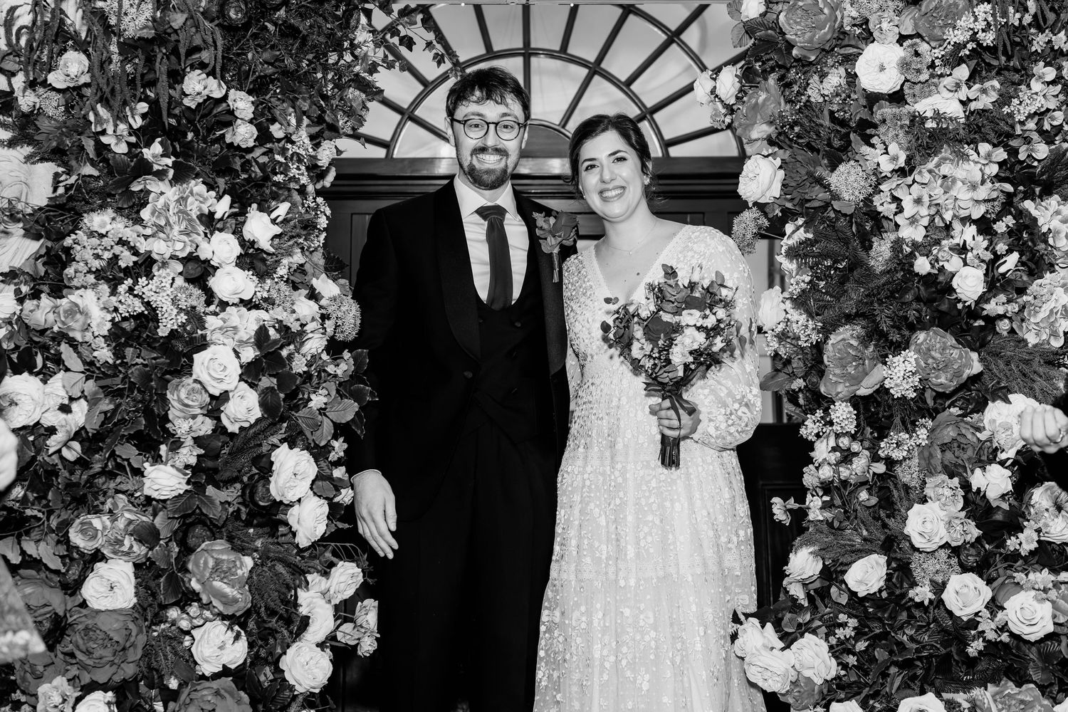 Black and white wedding photograph with couple standing together amid floral arrangements and decorative archways.