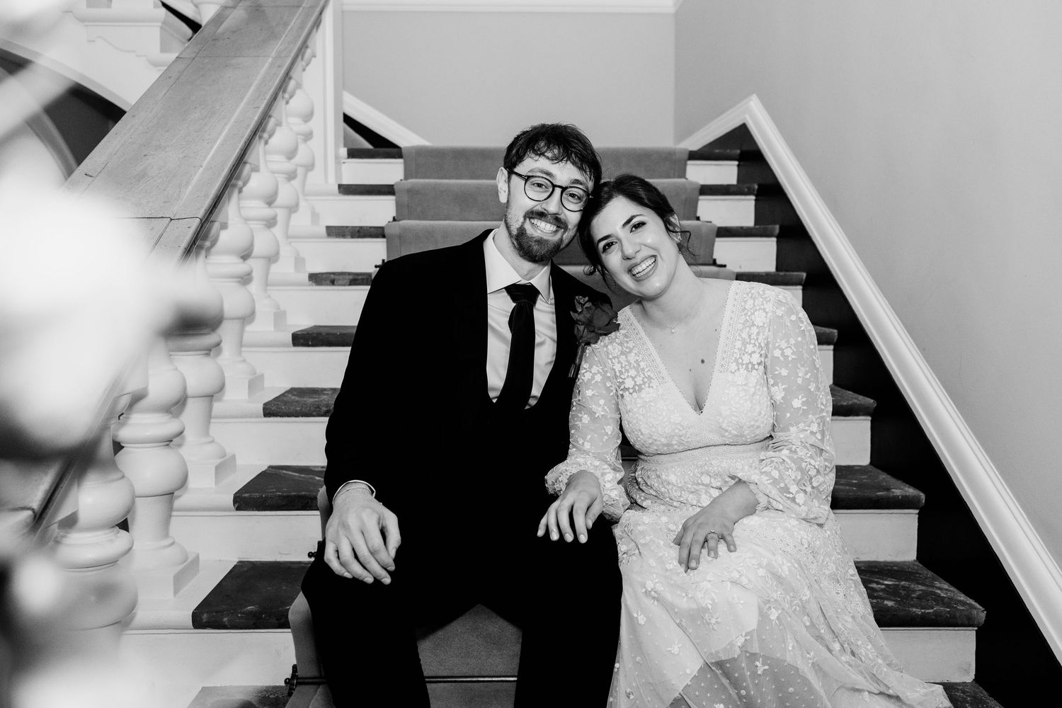 Black and white photo of a couple sitting together on white stairs, with the person in a wedding dress leaning in close.