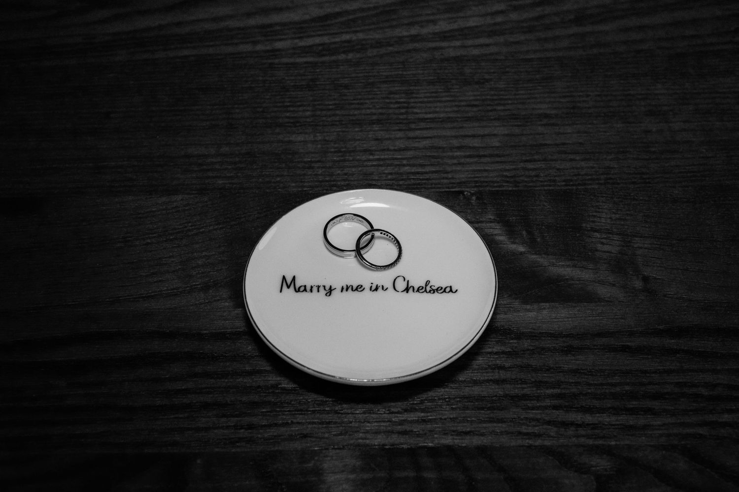 Two silver wedding rings sit on a white ceramic plate with handwritten text against a dark wooden background.