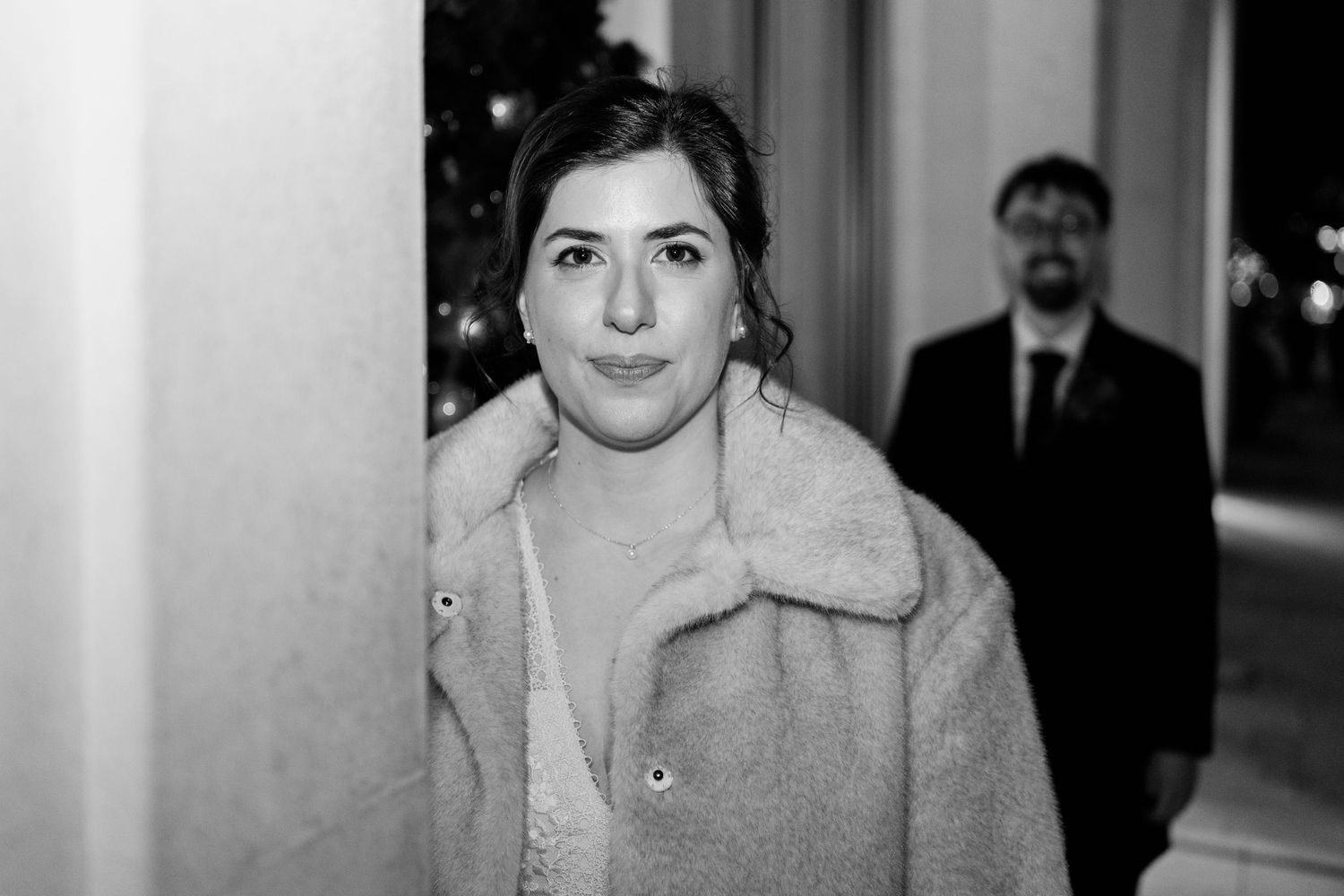 Black and white photo showing someone wearing a light colored coat with fur collar in an indoor hallway.