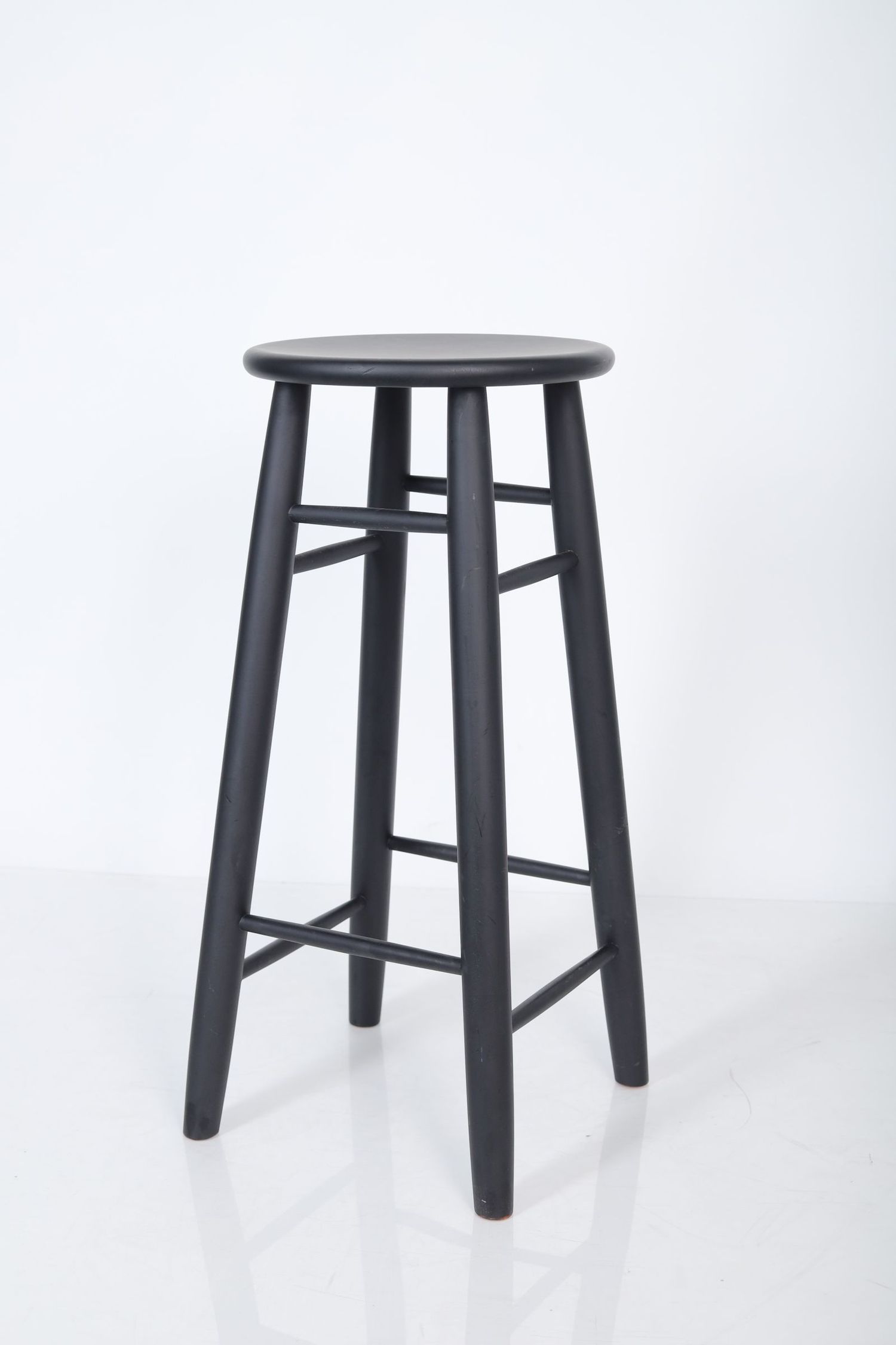 A tall black wooden bar stool with a round seat and four tapered legs stands against a white background.