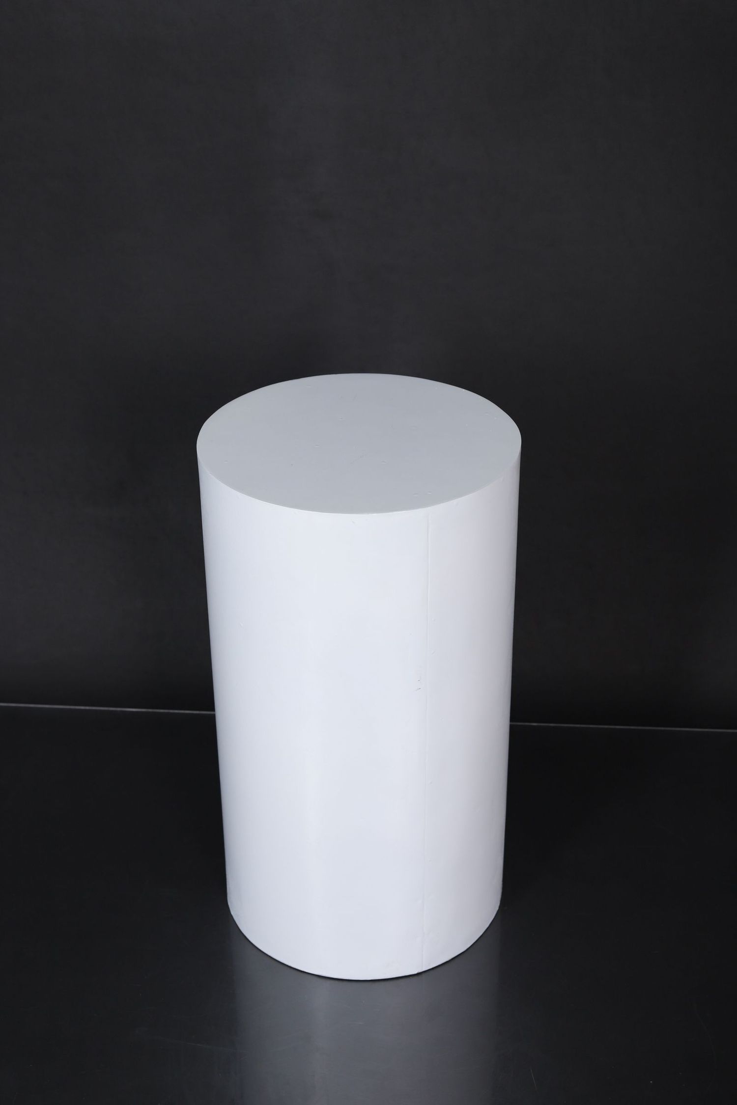 White cylindrical pedestal or display stand photographed against a black background in dramatic lighting.
