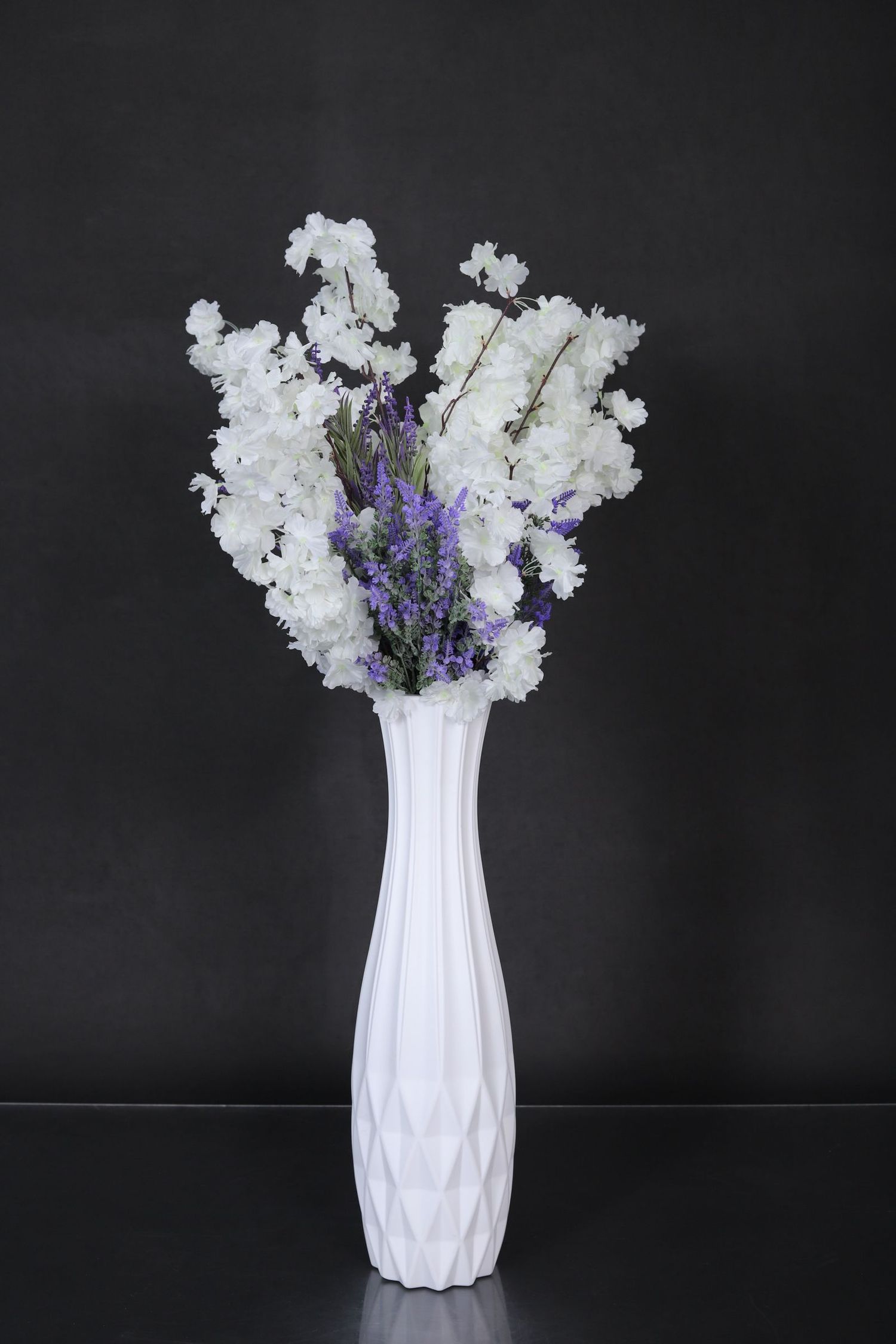 White and purple floral arrangement in a tall geometric white ceramic vase against a dark background.