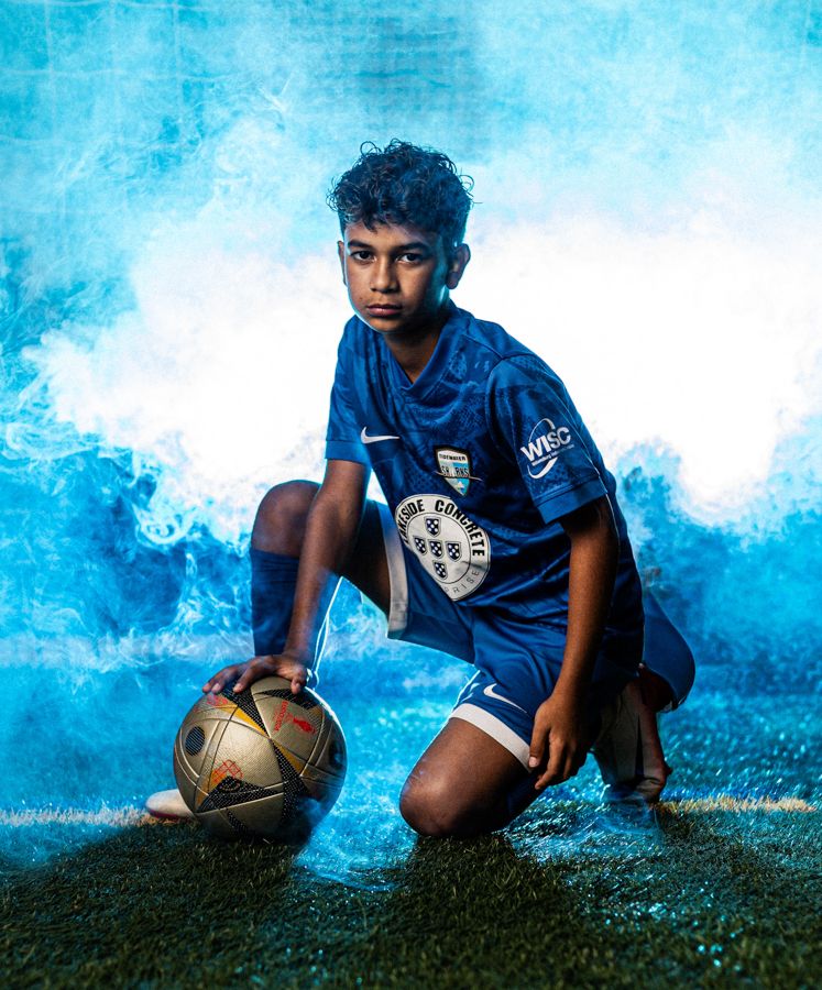 dramatic sports portraits in williamsburg virginia boy kneeling down on a soccer ball with blue smoke behind