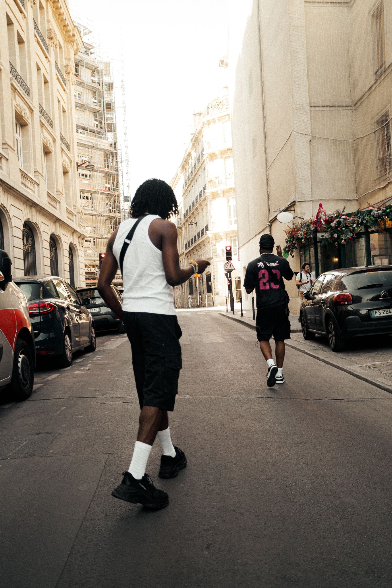 Two people walking down a narrow urban street lined with classic Parisian buildings and parked cars.