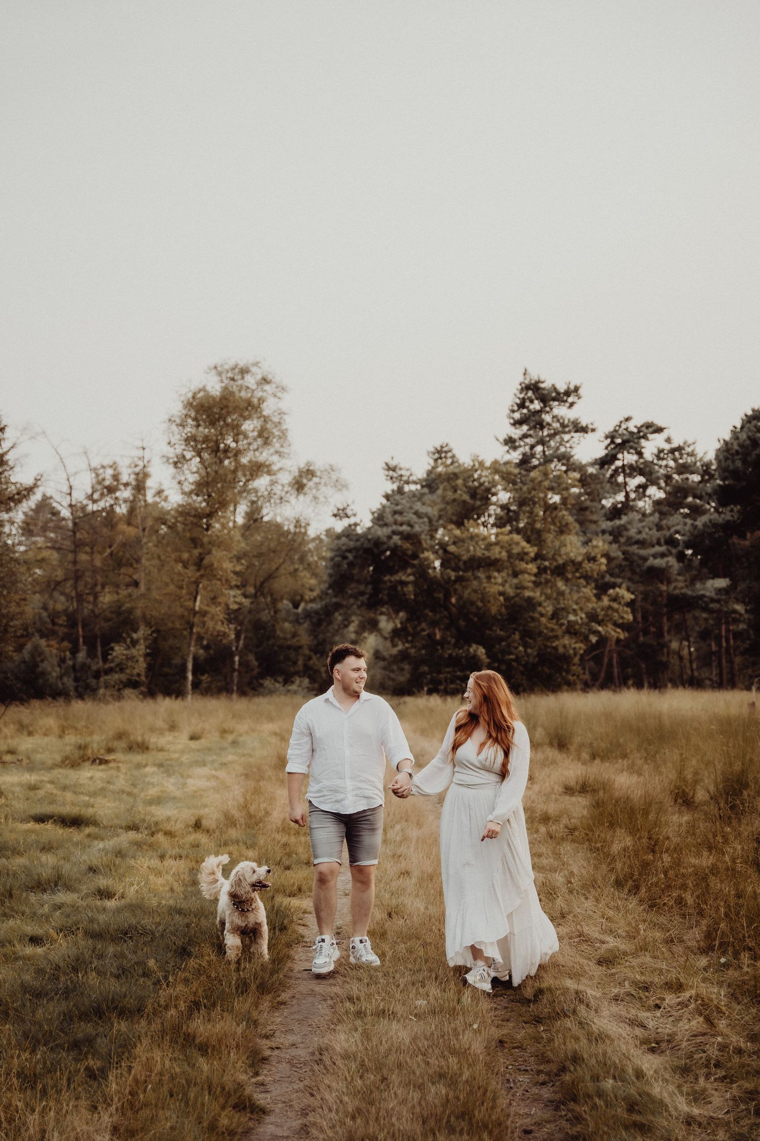 Couple walking with their dog on a rustic dirt path through a grassy field at golden hour.