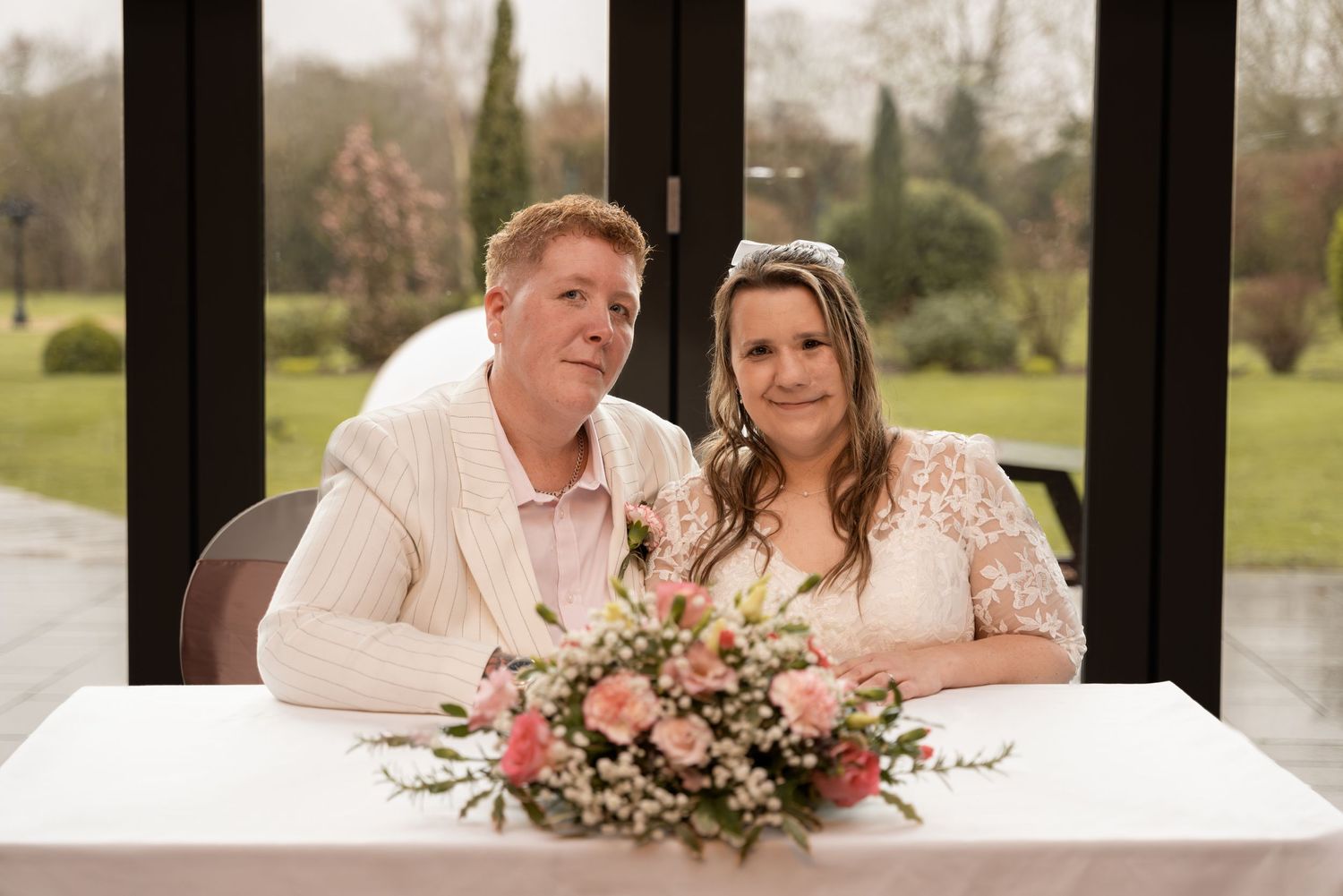 Happy couple sitting at a table signing wedding documents, with pink floral arrangement during a wedding in Lincolnshire.