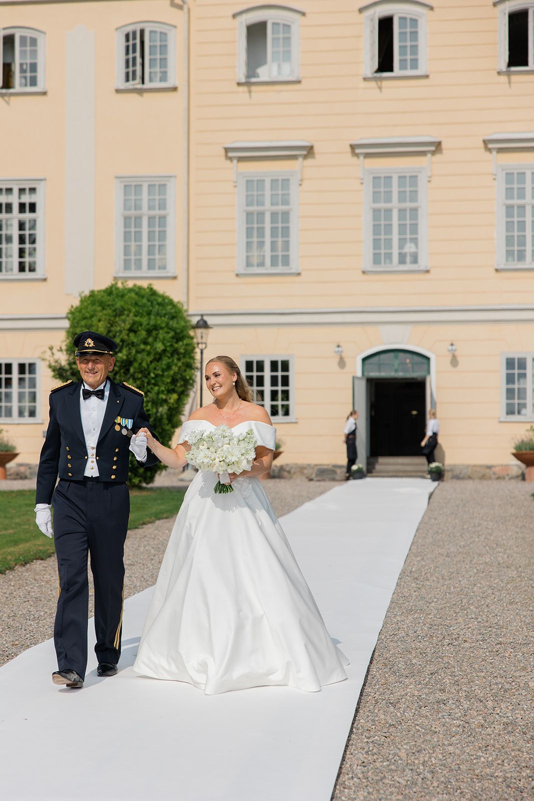 A couple in formal wedding attire walks along a gravel path outside an elegant cream-colored European manor house.