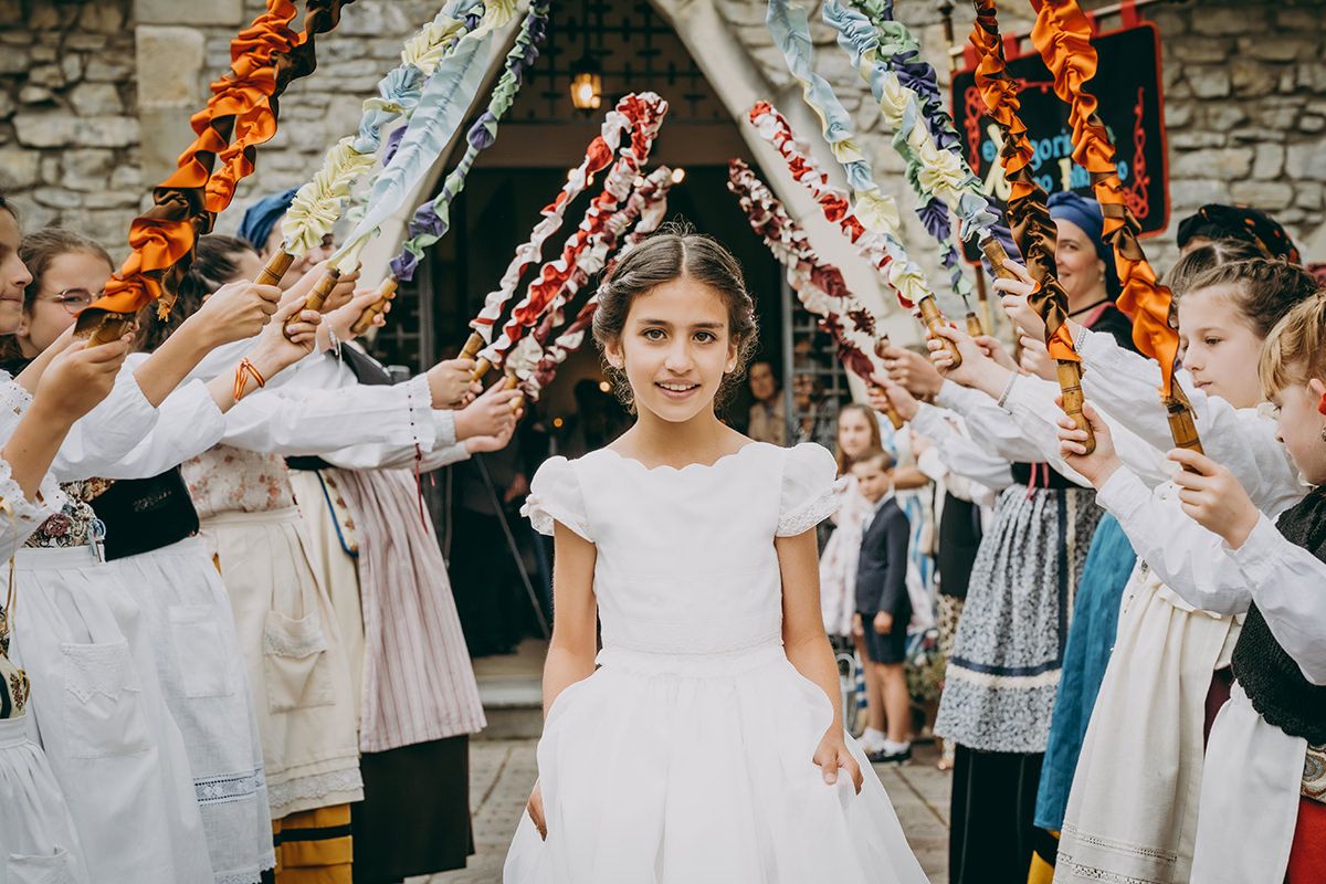 Niña de comunión saliendo de la iglesia entre un pasillo de niños con trajes tradicionales y varas decoradas en Asturias.