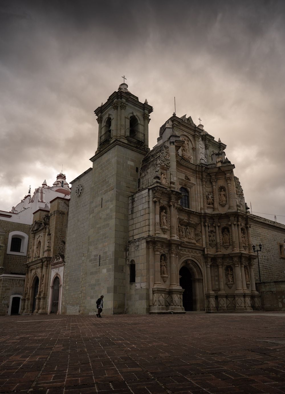 Historic stone cathedral facade with ornate baroque architecture photographed under dramatic cloudy skies in Oaxaca, Mexico.