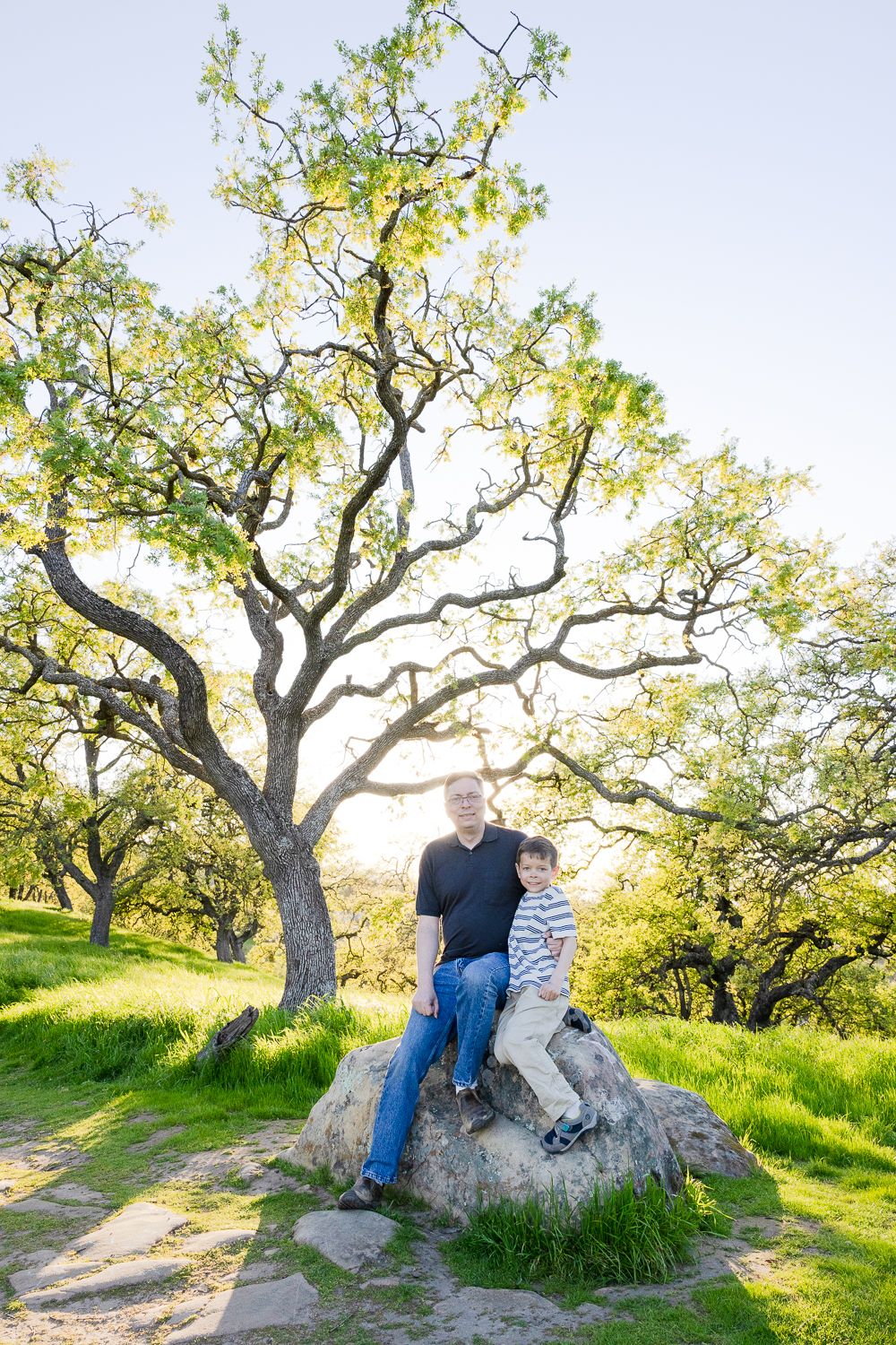 A father and son sit together on a large rock under sprawling oak trees in a sunlit grassy meadow in San Jose at sunset.