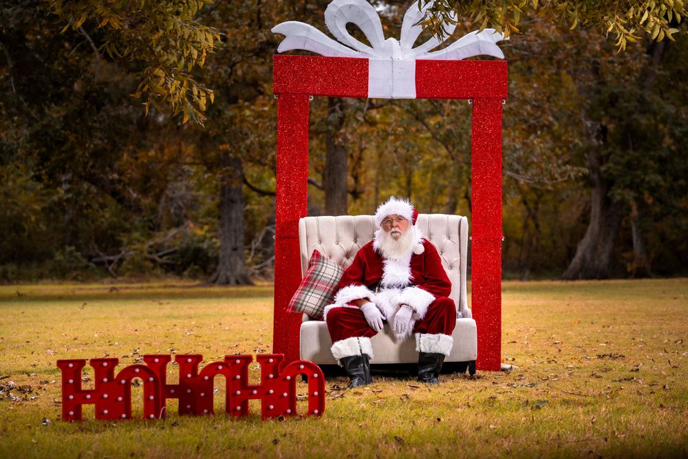 Santa seated in large red gift box display outdoors with ho ho ho decorations.