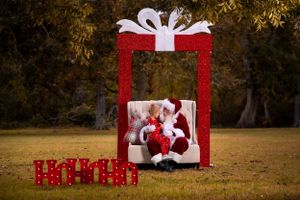 A large red glittering gift box display with a white bow sits on grass with holiday decorations beneath it.