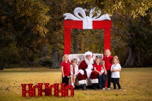 Red and white holiday photo setup outdoors with large present frame and decorative boxes against autumn trees.