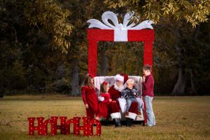A large red present photo backdrop with figures in red holiday outfits sitting beneath a white bow outdoors.