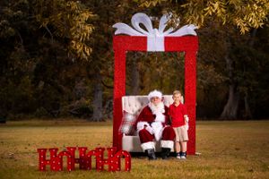A festive outdoor Christmas scene with a large red gift box prop and decorative presents against a golden autumn backdrop.