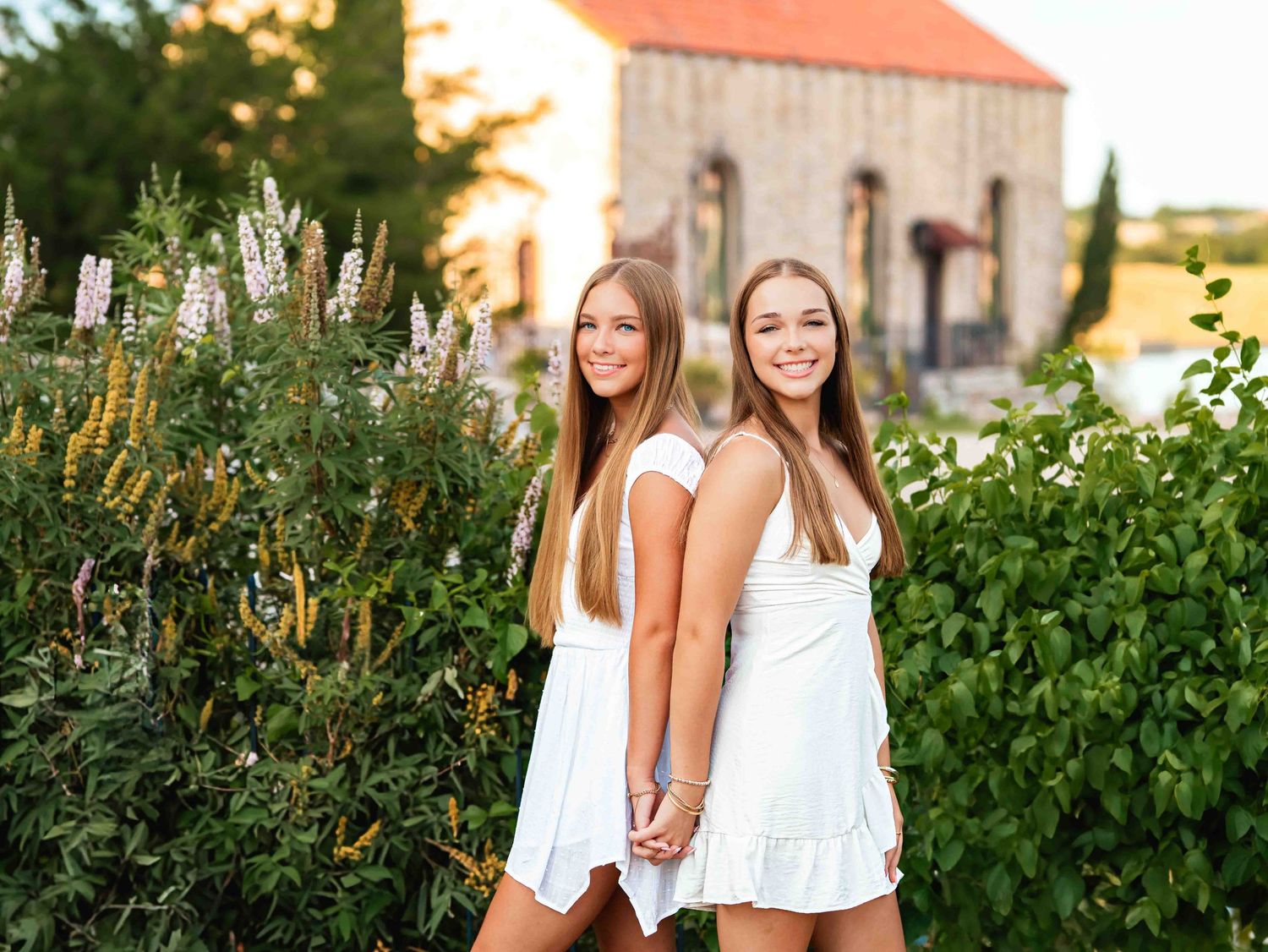 Two people in white dresses stand together outdoors with green foliage and warm sunset lighting in the background.