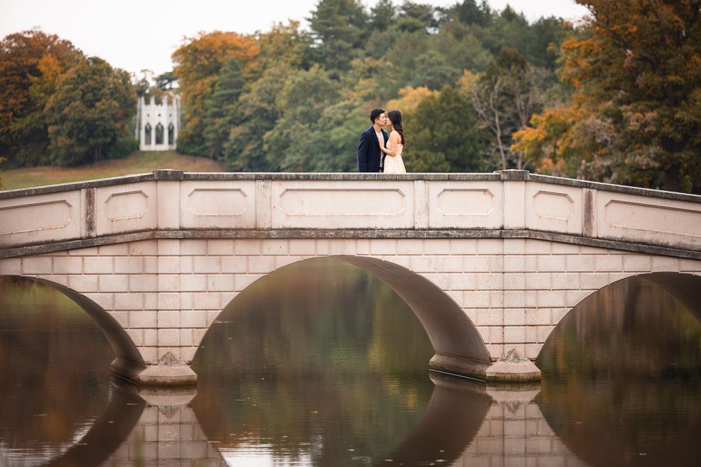 An engagement shoot in Painshill Park, Surrey. A newly engaged couple share an embrace on a stone bridge over the lake.