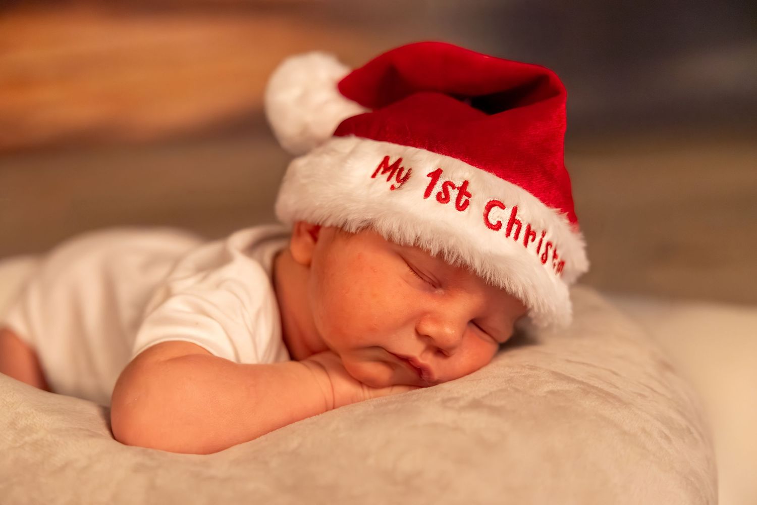 A sleeping newborn baby wearing a red and white "My First Christmas" Santa hat rests peacefully on soft fabric.