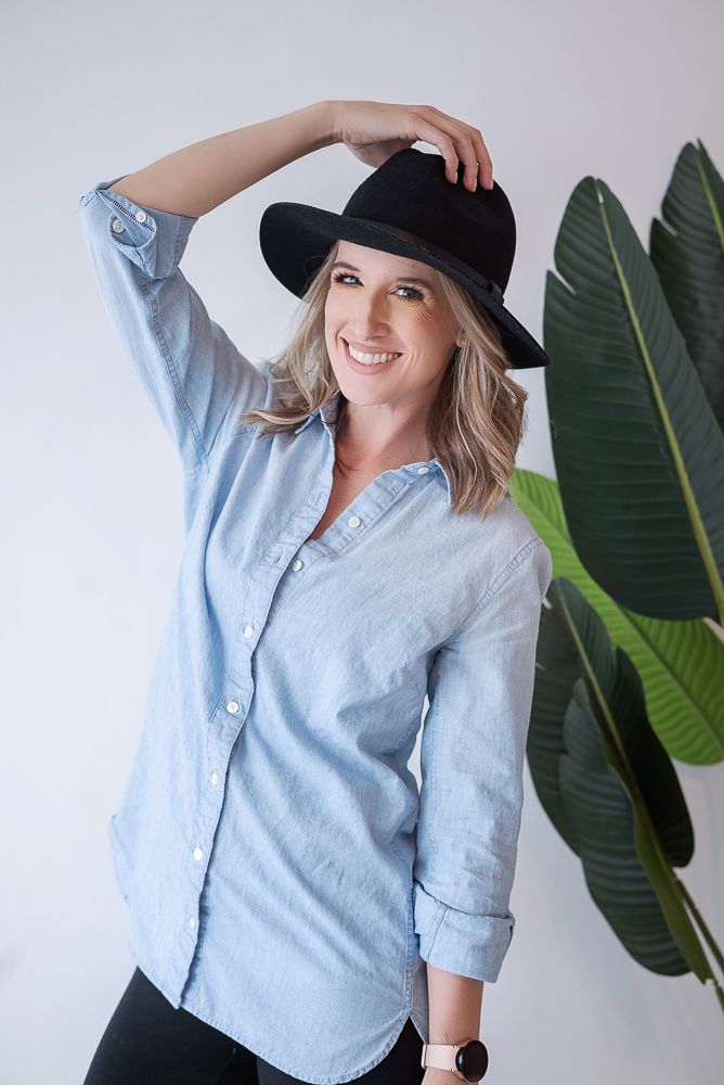 Woman in light blue button-down shirt and black hat poses playfully against white wall with large green plant leaves.