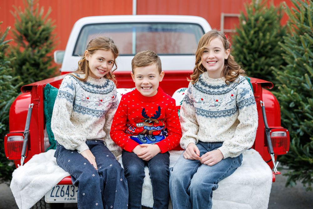 Three siblings in winter sweaters sitting in the bed of a red pickup truck surrounded by Christmas trees.