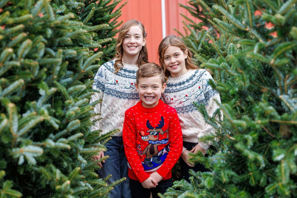 Three children in coordinated winter sweaters pose among evergreen trees with a red barn background.