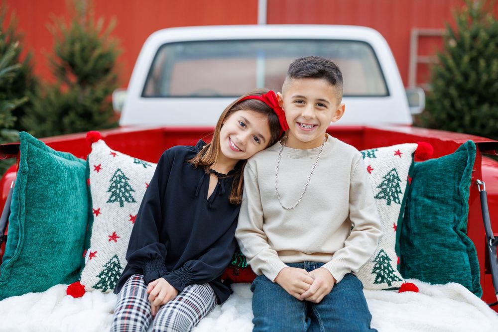 Two children sitting in truck bed with festive Christmas pillows and red pickup truck background.