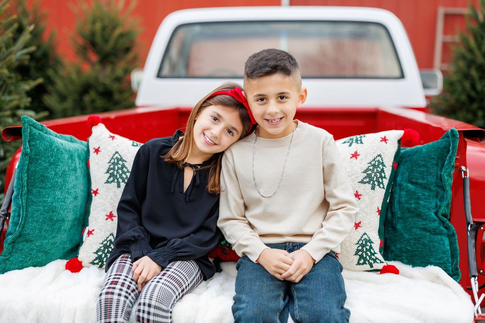 Two children sitting together in red truck bed with green Christmas pillows.