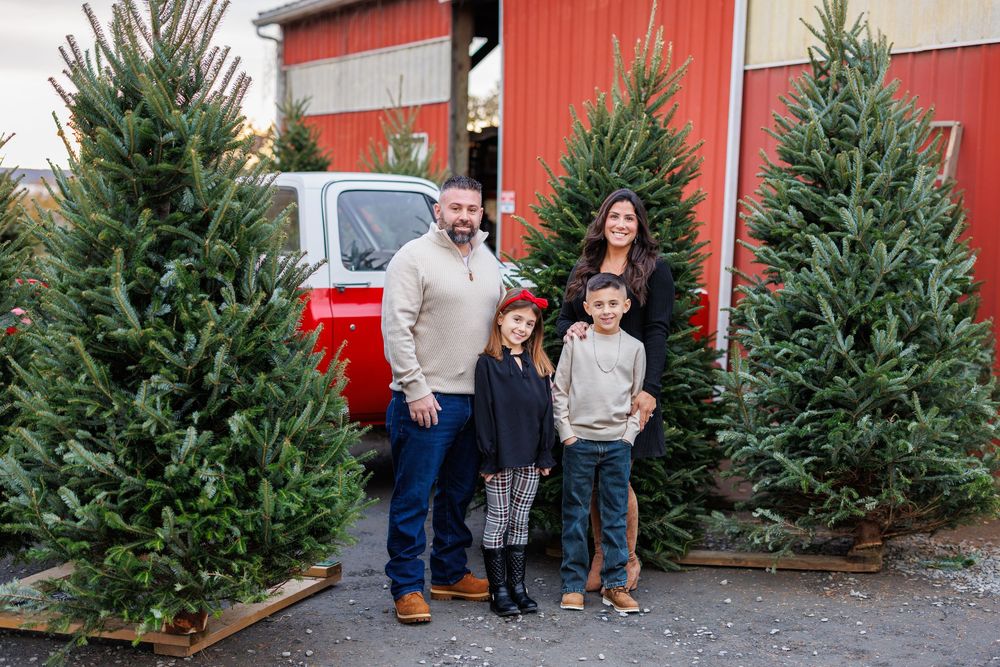 Family of four standing together at a Christmas tree farm with red barn and evergreens.