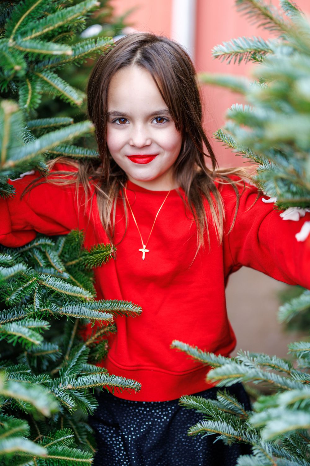 A child in a red sweater poses among evergreen branches during holiday photos.