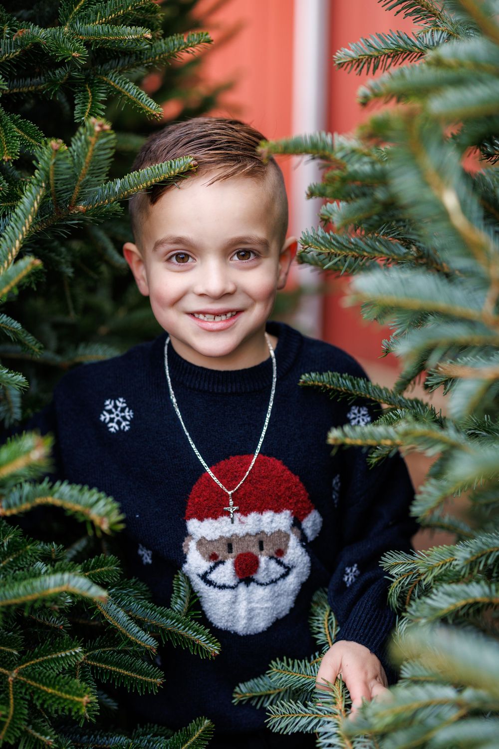 A child wearing a festive Santa sweater smiles among Christmas tree branches.