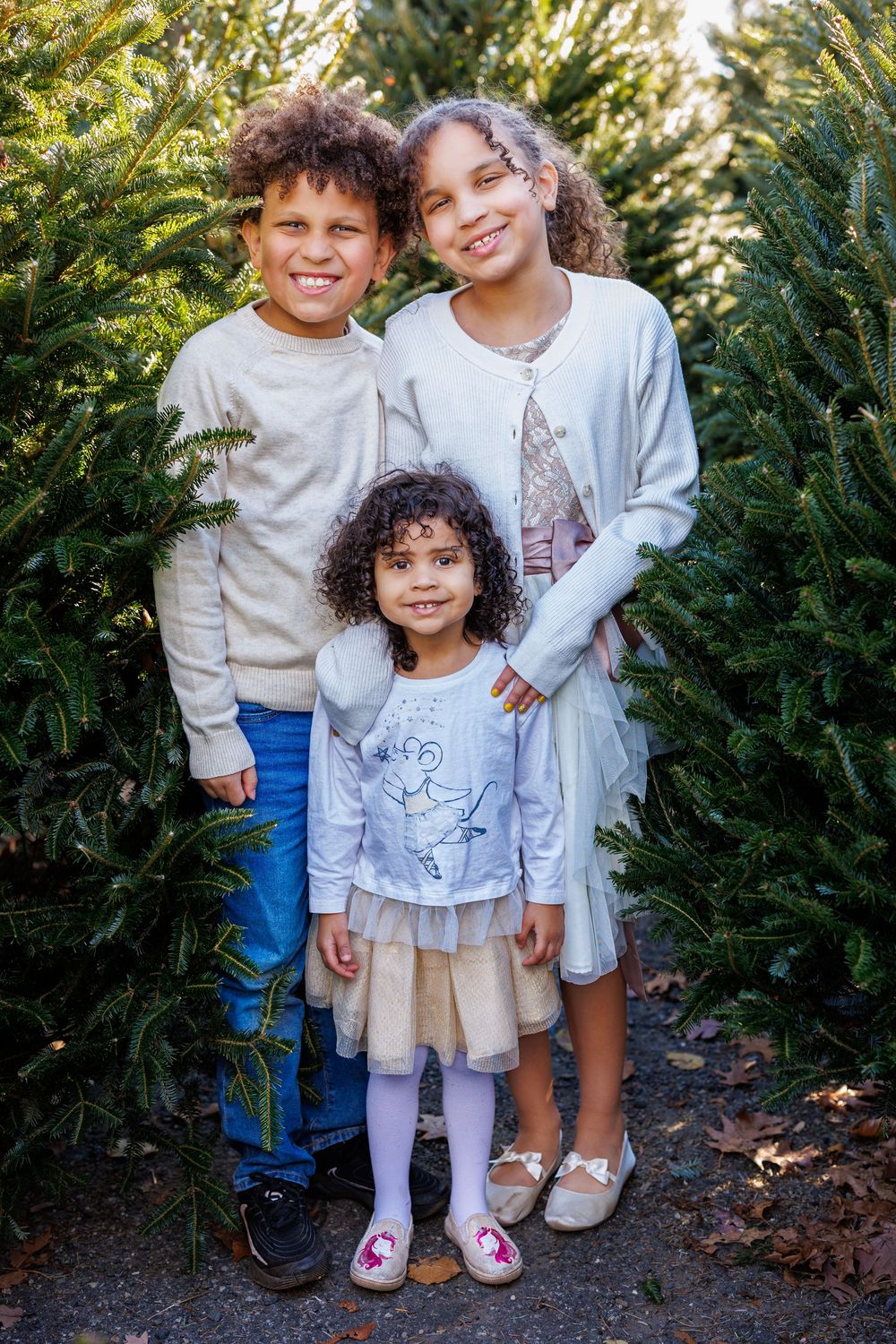 Three children standing together by evergreen trees wearing casual clothing.