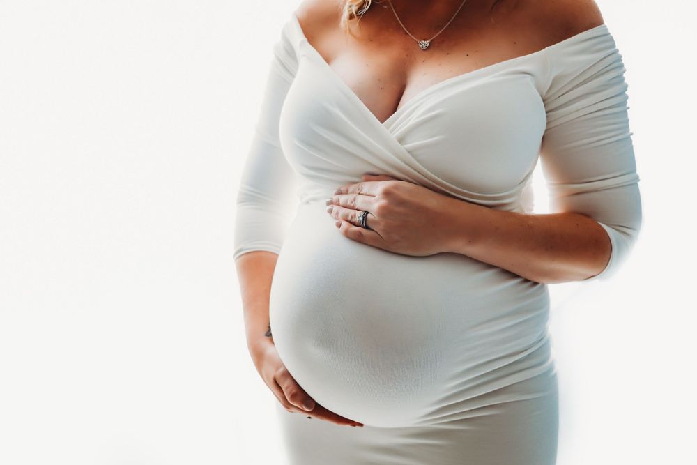 Maternity photos showing pregnant woman in white dress against white background.