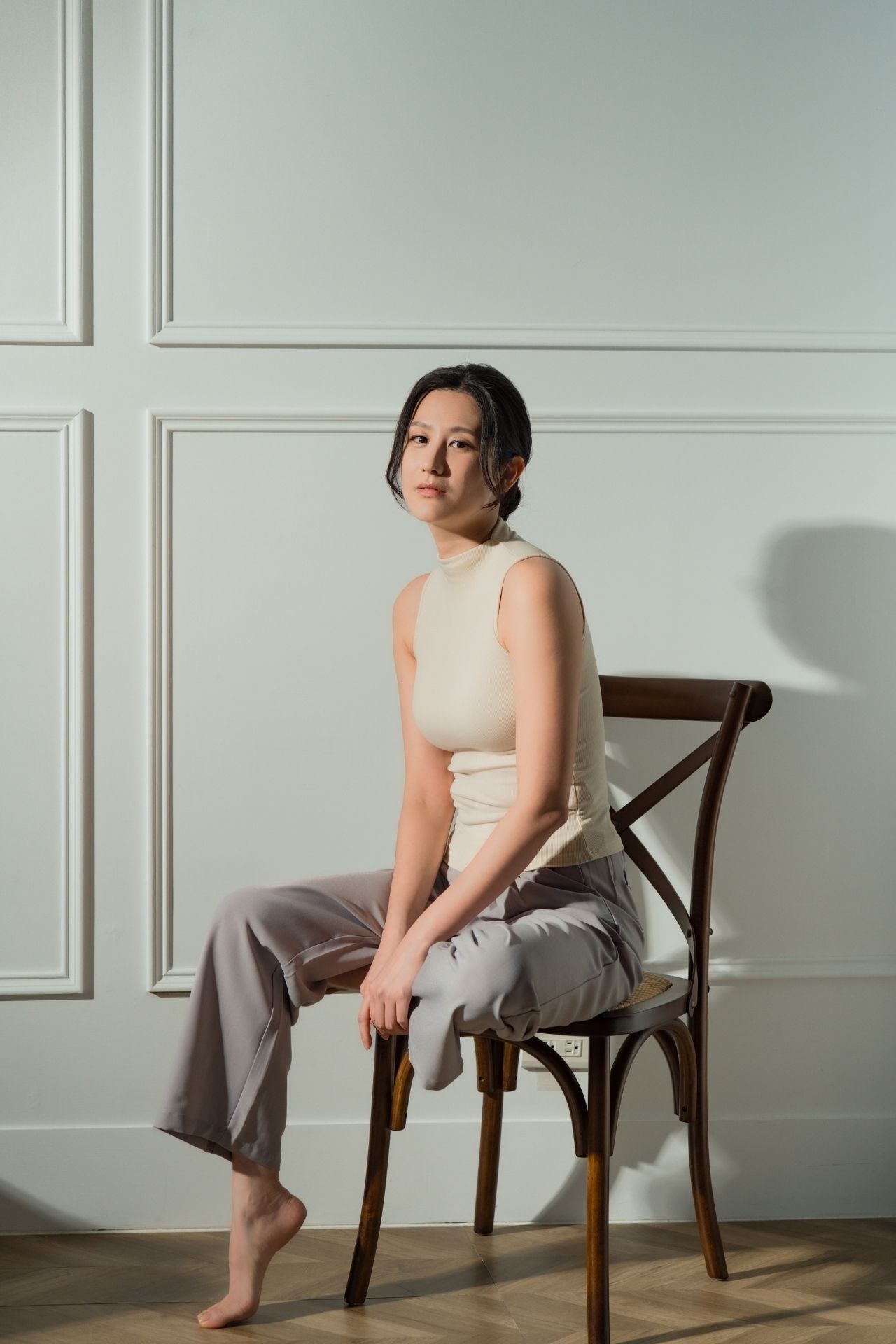 A person sits in various poses on a wooden chair against a white wall in a minimalist setting.
