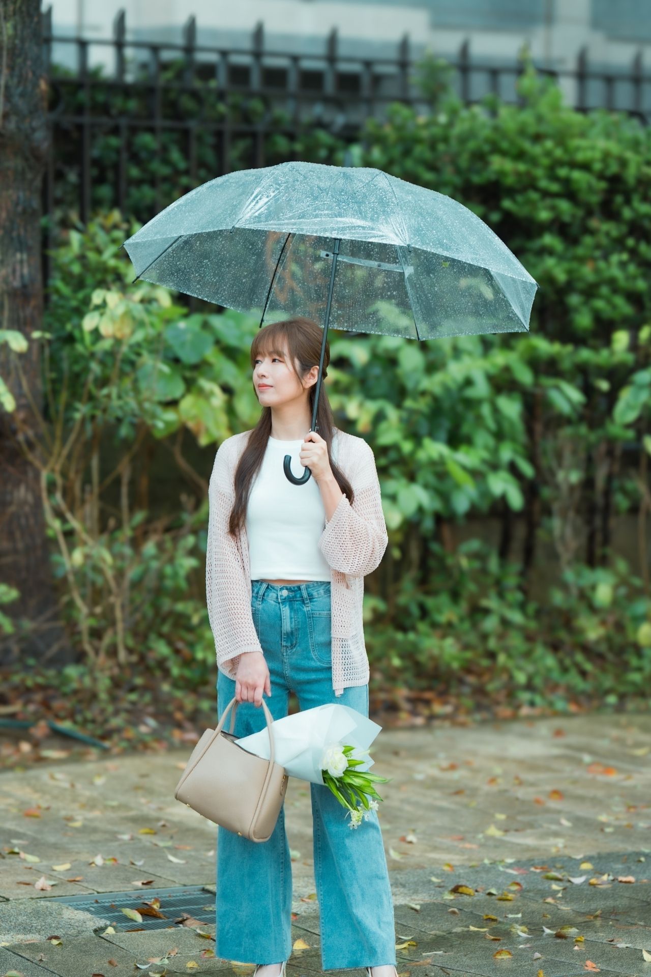A person in a teal jumpsuit and white sweater holds a clear umbrella while walking through a garden path on a rainy day.