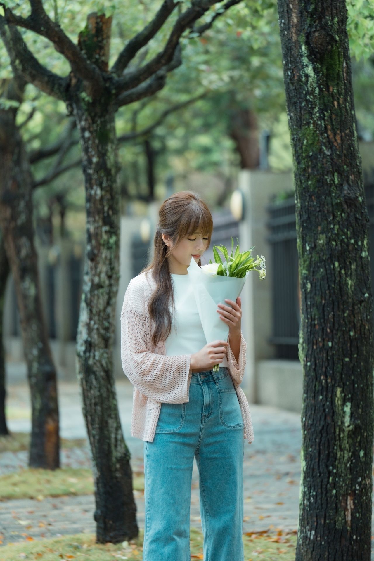 Someone in a pink cardigan and blue jeans holds fresh produce while walking along a tree-lined path.