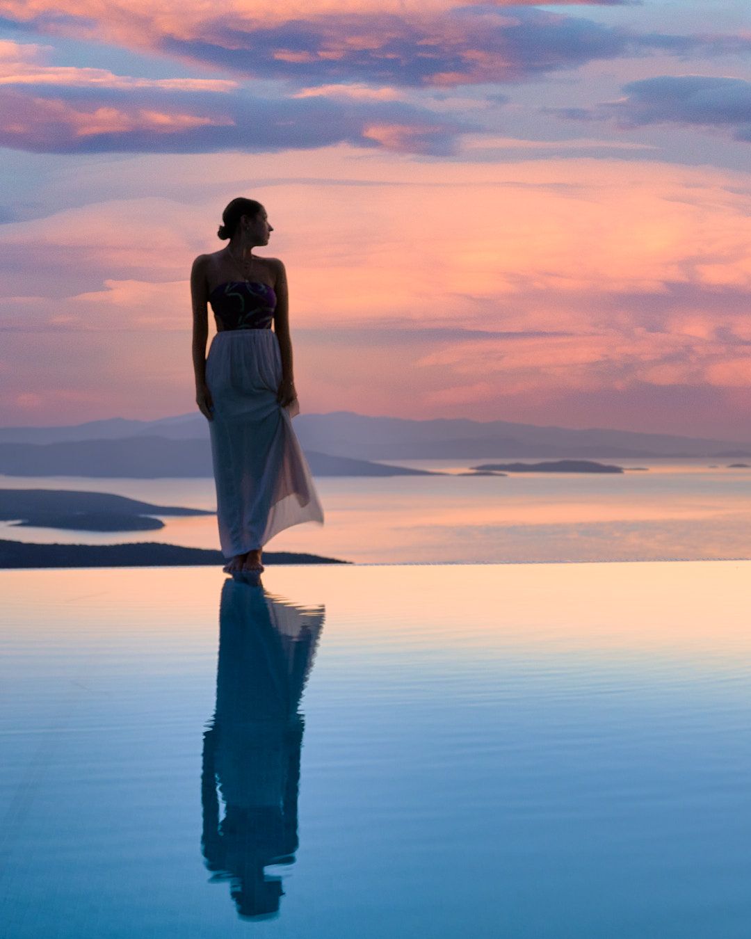 Woman standing near the sea at sunset, reflected on calm water with soft blue and pink tones.